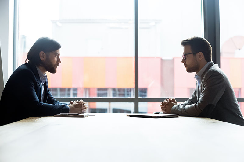 Two men sit across from each other at a table in an office setting, facing each other with serious expressions, hands clasped, and large windows in the background letting in natural light.