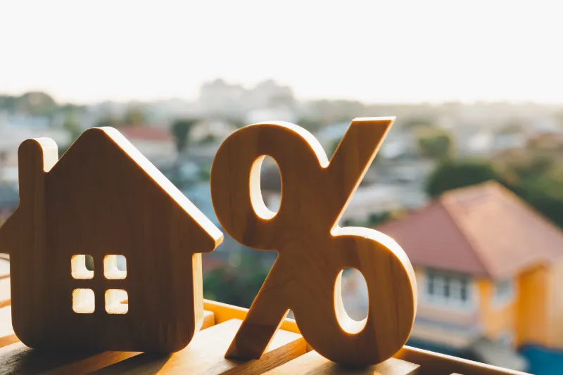 Wooden house and percentage symbol figures on a table with blurred houses and sunlight in the background, representing mortgage or interest rates in real estate.