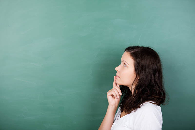 A woman with long brown hair, wearing a white shirt, stands in profile against a green chalkboard, looking thoughtful with her finger resting on her chin.