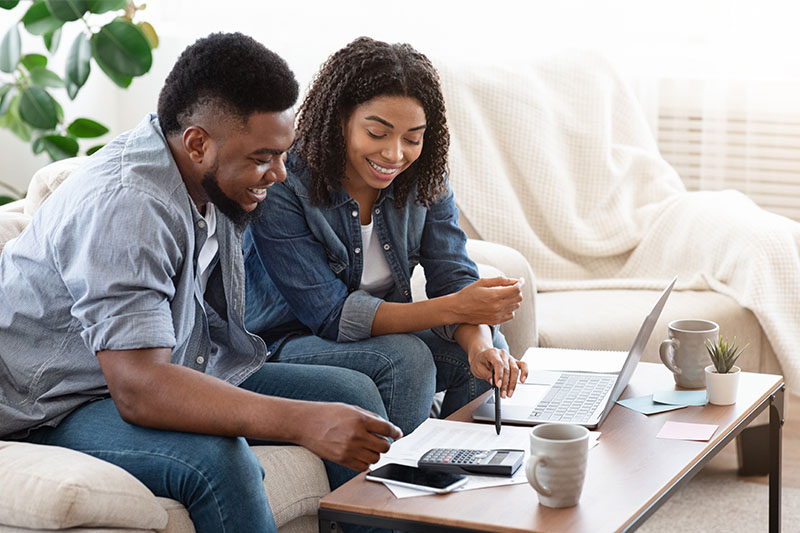 A smiling couple sits on a couch, reviewing financial documents and using a laptop and calculator on a coffee table, appearing focused and happy while managing their finances at home.