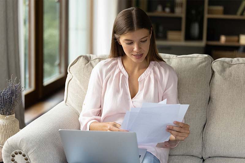 A woman sits on a couch with a laptop, looking at papers in her hands. She wears a light pink blouse and appears to be reviewing documents in a bright, cozy living room.