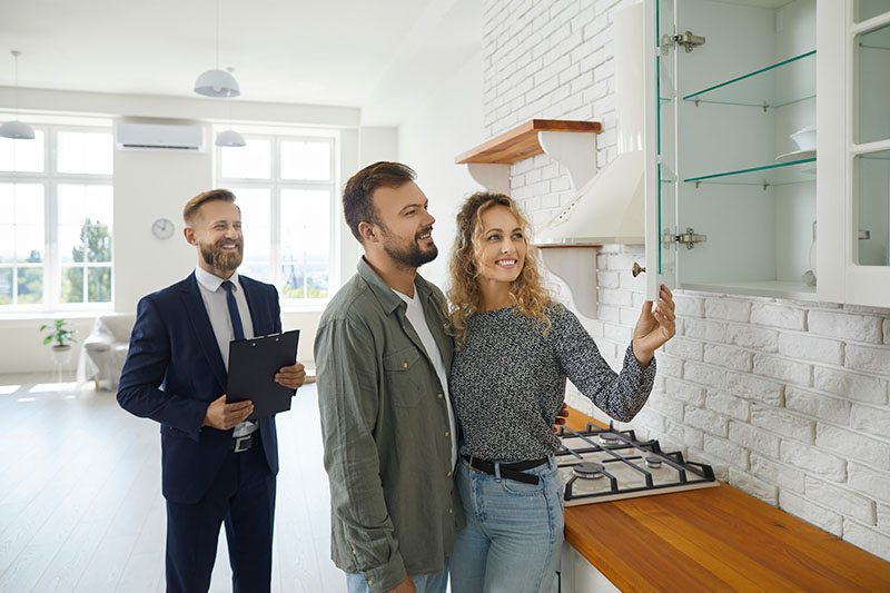 A smiling couple examines a glass kitchen cabinet in a modern, bright kitchen while a real estate agent in a suit stands nearby holding a clipboard.