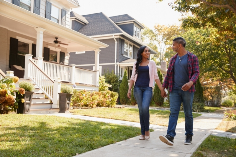 A couple walks hand in hand on a sidewalk in a suburban neighborhood, passing by houses with front porches and gardens on a sunny day.