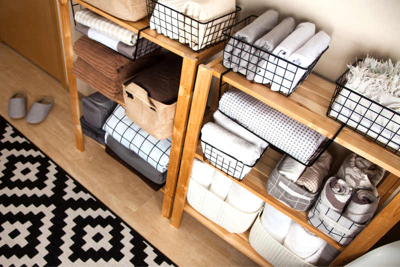 Wooden shelves neatly organize towels, linens, and baskets in a bright room. A black-and-white geometric rug and gray slippers are on the floor, creating a tidy, cozy atmosphere.