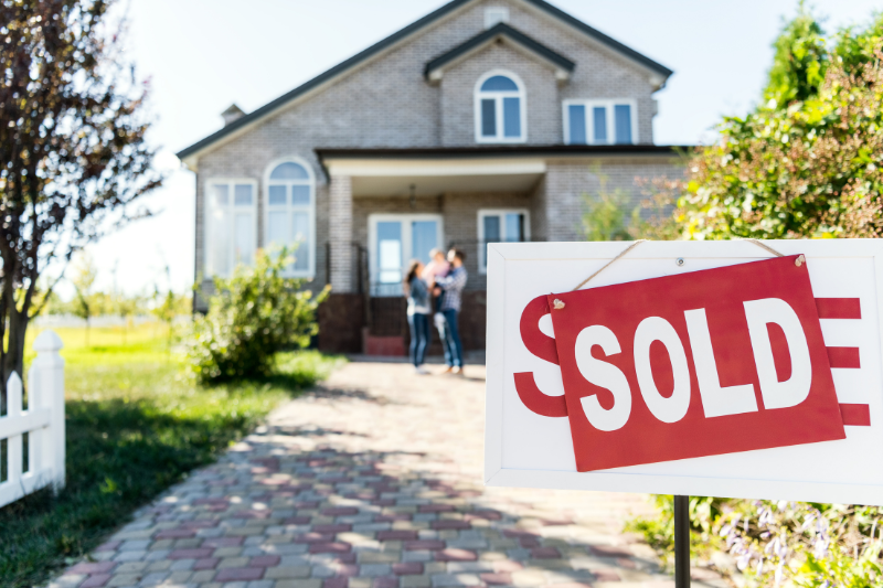 A house with a SOLD sign in front, with a blurred family standing together near the entrance, suggesting they are the new homeowners. The scene is bright and cheerful.