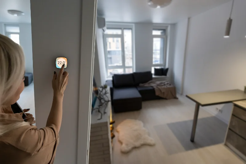 A person adjusts a smart thermostat on a wall, setting it to 22°C in a modern, bright living room with large windows, a dark sofa, a table, and light-colored flooring.
