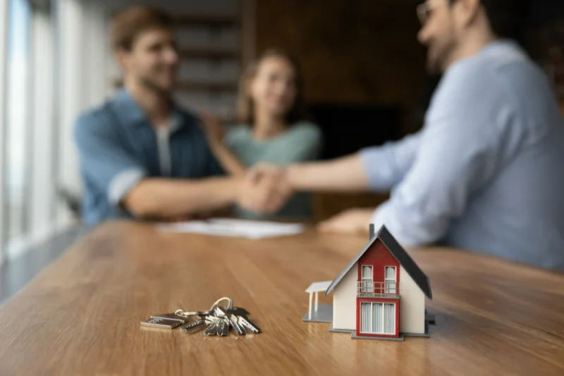 A miniature house and keys sit on a wooden table in focus, while two people shake hands and a third person looks on in the blurred background, suggesting a real estate transaction.