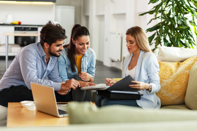 Three people sit on a sofa in a bright living room, looking at documents together. A woman holds a folder, while a man and another woman listen and discuss. A laptop and coffee cup are on the table in front of them.