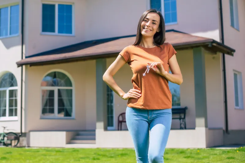 A smiling woman stands in front of a house, holding a set of keys in her hand, suggesting she is a new homeowner. She is wearing a rust-colored T-shirt and blue jeans.