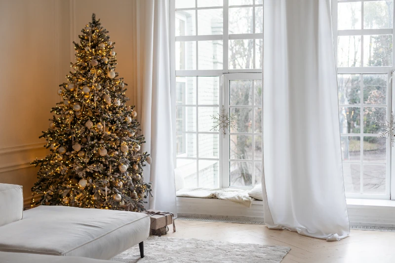 A decorated Christmas tree with white and gold ornaments and lights stands in a bright living room next to large windows with white curtains. A few wrapped gifts sit beneath the tree.