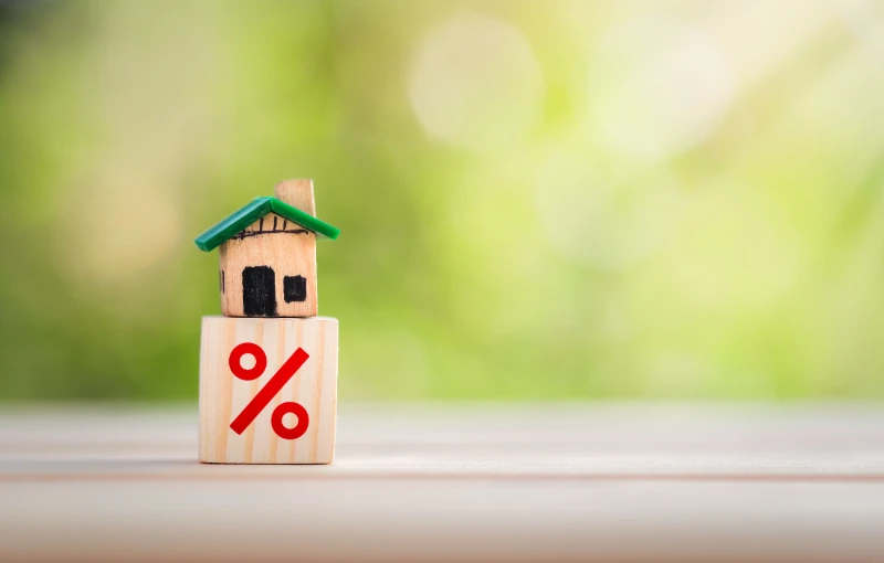 A small wooden house figurine sits on top of a wooden block with a red percent symbol, against a blurred green and beige background.