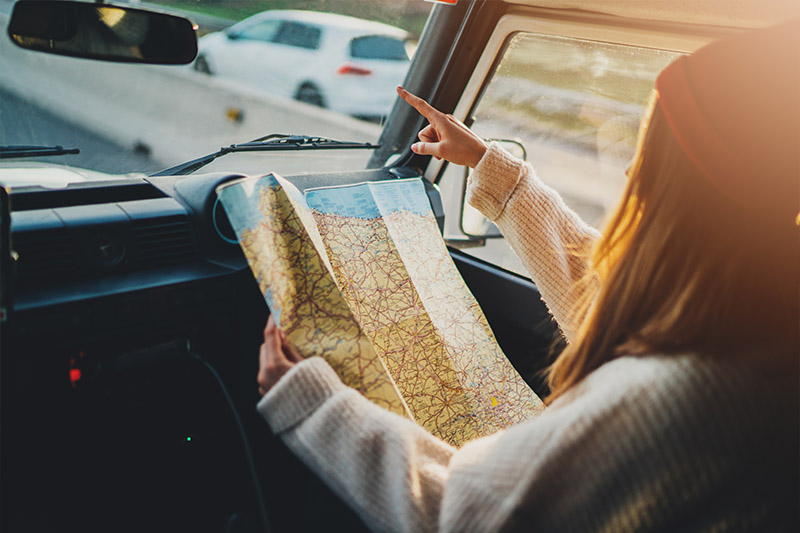 Learning Center 7 A person in a sweater and hat sits in a car passenger seat, holding a map while pointing out the windshield, suggesting they are navigating or giving directions during a road trip. Sunlight streams through the window.