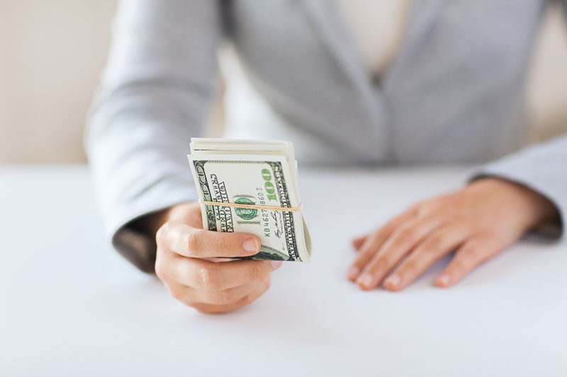 Learning Center 3 A person in a light gray suit holds out a stack of U.S. hundred-dollar bills secured with a rubber band, while their other hand rests on a white table.