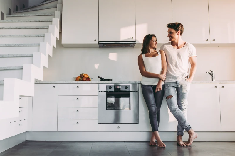 Seller-Paid Buydown 1 A young couple, both casually dressed in white tops and jeans, stands barefoot in a modern white kitchen, smiling at each other while leaning against the counter near a staircase.