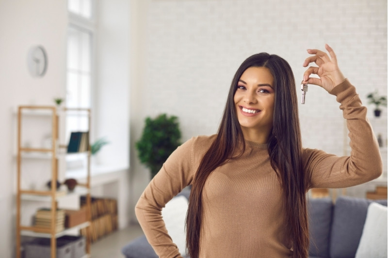A smiling woman with long brown hair stands in a bright living room, holding up a key in her right hand, suggesting she has just moved in or bought a new home.