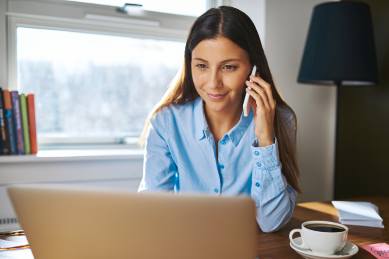 A woman in a blue shirt sits at a desk, talking on her phone while looking at a laptop. There is a cup of coffee, some papers, and a lamp nearby, with sunlight coming through the window behind her.