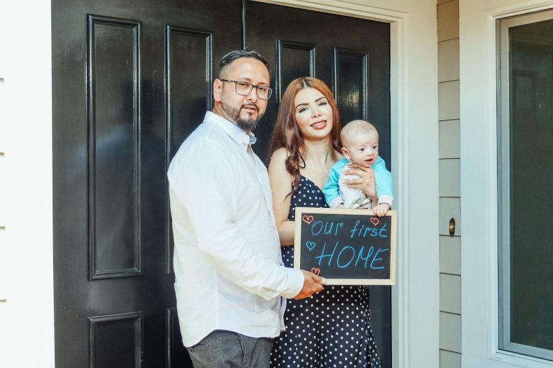 Young family with baby holding a sign Out First Home at door