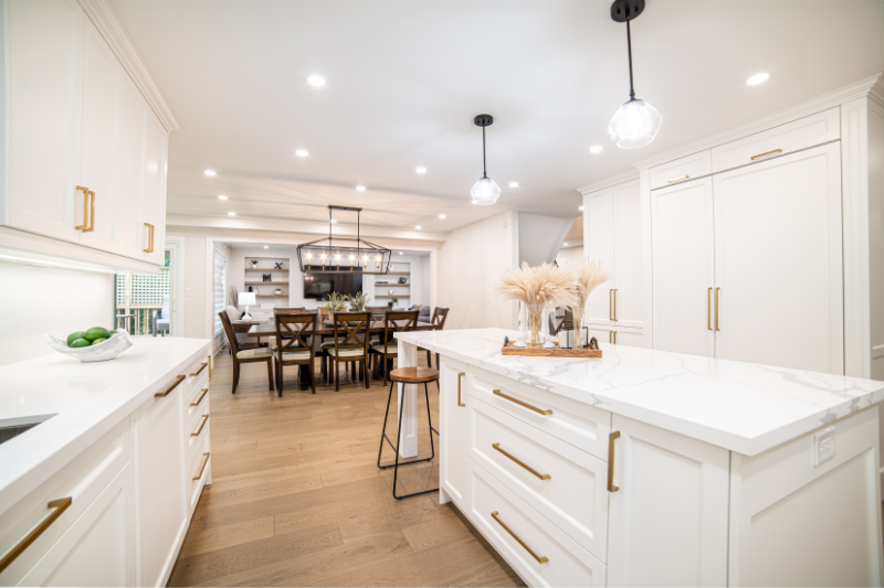 Spacious modern kitchen with white cabinetry, gold hardware, marble countertops, wooden floors, and pendant lights. A kitchen island with stools and decorative accents faces a dining area and living space in the background.