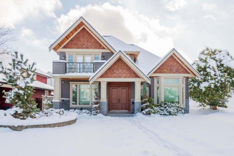 A modern two-story house with wood and brick accents surrounded by snow, trees, and bushes. The sky is partly cloudy and the house has large windows, a balcony, and a double front door.