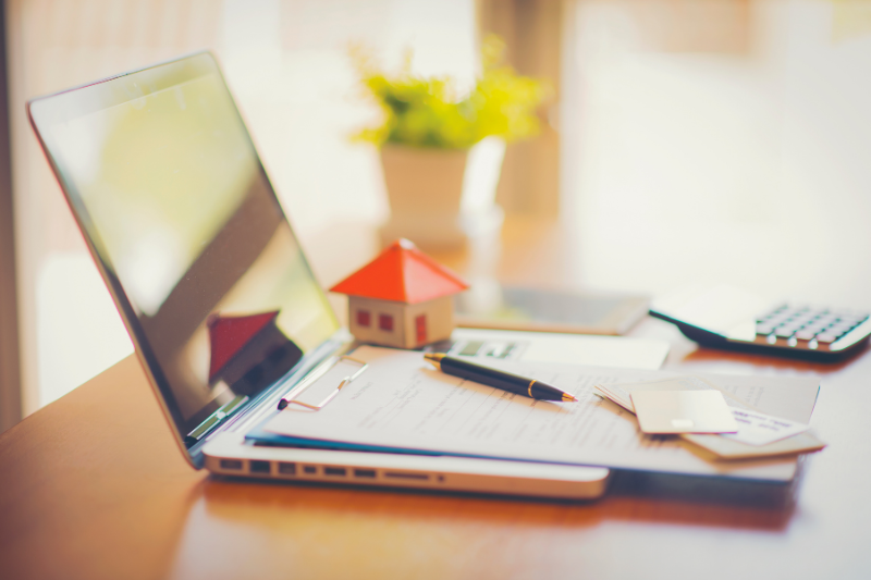 A laptop, paperwork, a pen, credit cards, a small model house, and a calculator are arranged on a desk with a potted plant in the background, suggesting a work-from-home or real estate theme.