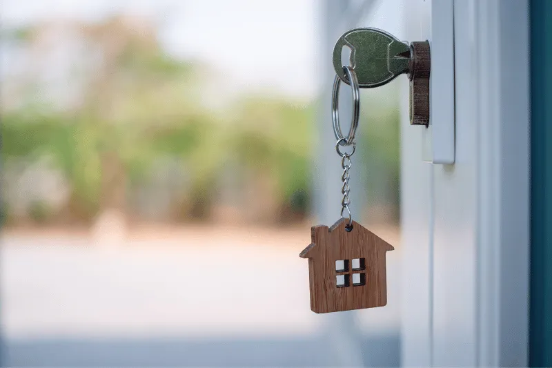 A silver key with a wooden house-shaped keychain is inserted into a door lock, with an outdoor background softly blurred.