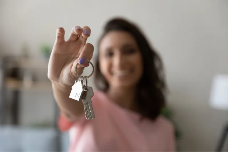 A smiling woman holds a set of keys with a house-shaped keychain toward the camera, suggesting new home ownership. The background is softly blurred.