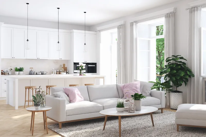 A bright, modern living room with white sofas, light wood furniture, green plants, and large windows letting in natural light. An open kitchen with white cabinets and pendant lights is visible in the background.