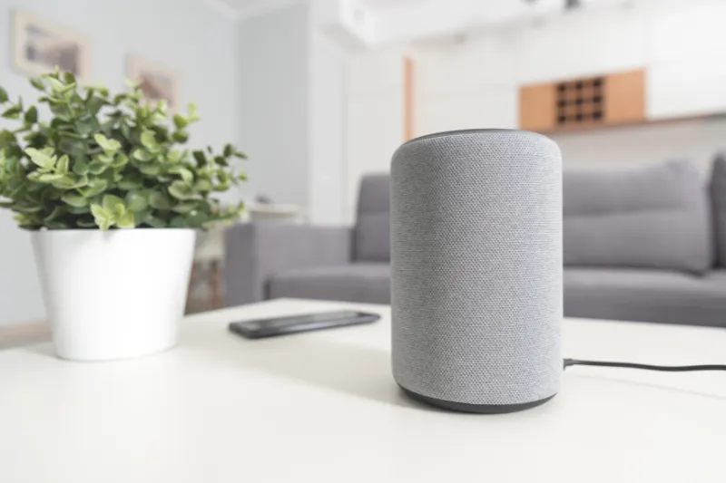 A gray smart speaker sits on a white table next to a potted green plant and a smartphone, with a sofa and shelves visible in the blurred background of a modern living room.