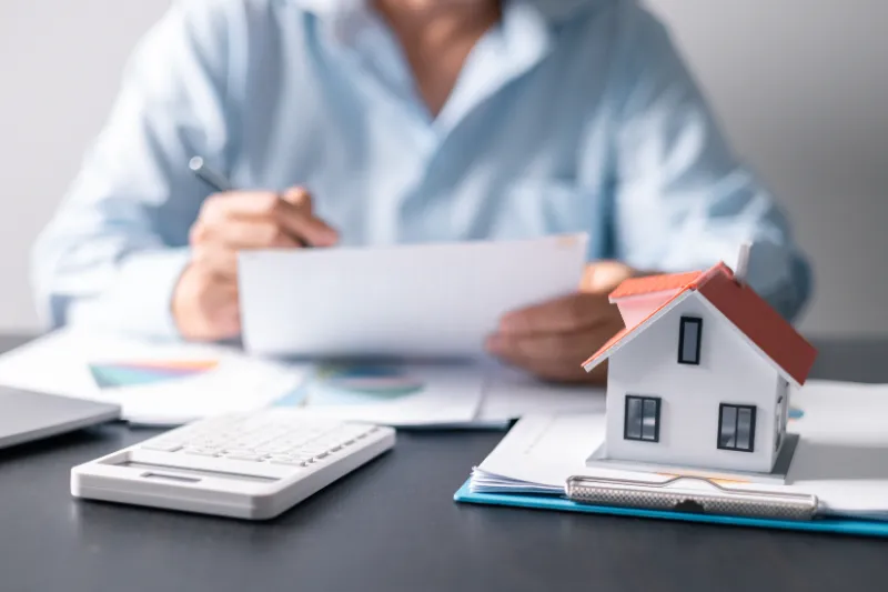 A person in a blue shirt reviews documents at a desk with a calculator, paperwork, pen, and a small model house, suggesting real estate, mortgage, or financial planning.