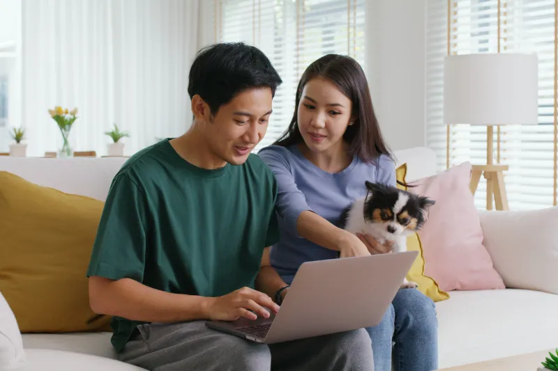 A man using a laptop sits on a sofa next to a woman holding a small dog. The woman points at the laptop screen. They appear to be discussing something together in a bright, modern living room.