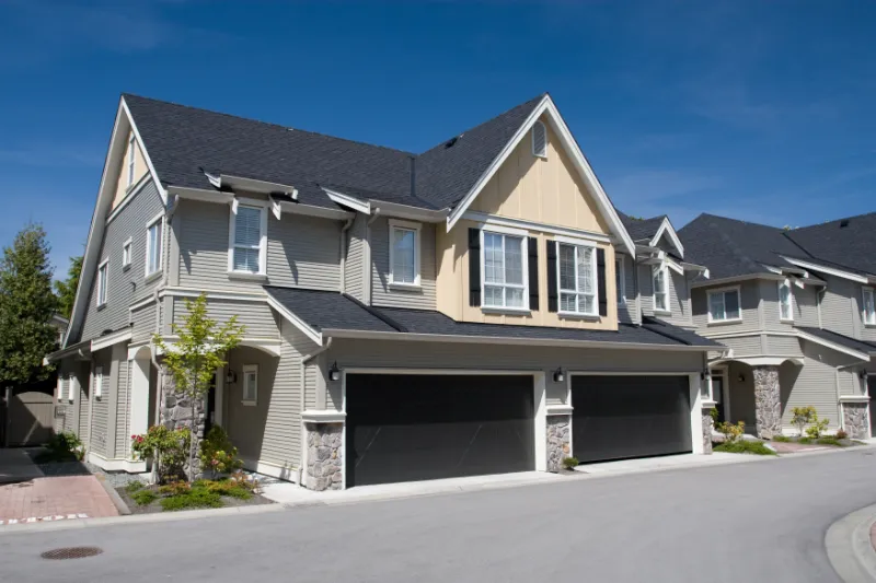 Two modern townhouses with beige siding, large windows, and black double garages are side by side on a sunny day, with a clean driveway and small trees in front.