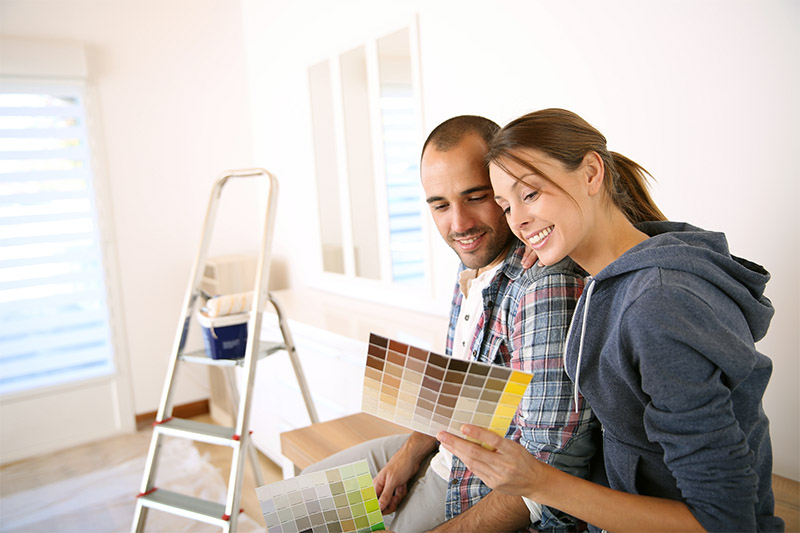 A smiling couple sits in a bright room, looking at paint color swatches. A ladder and painting supplies are in the background, suggesting they are planning to redecorate or paint the room together.