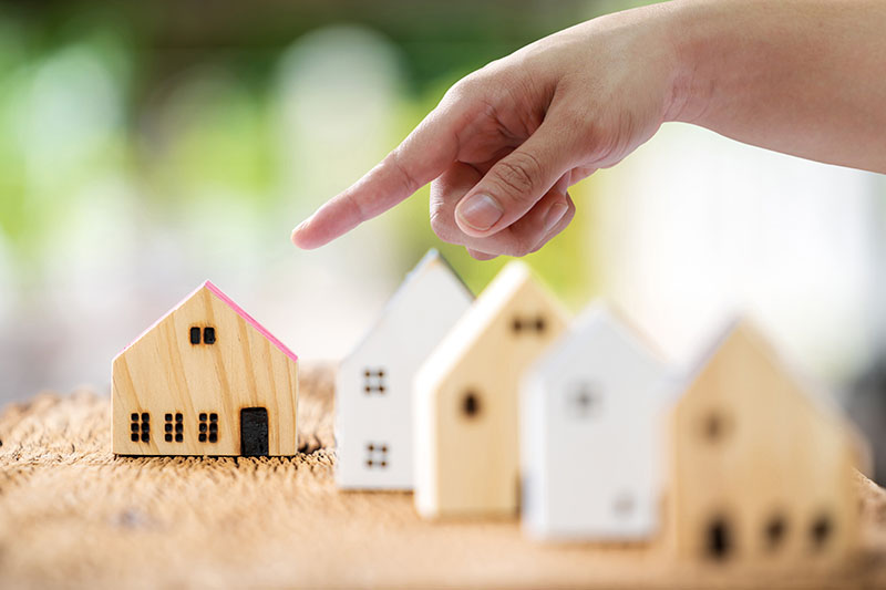 Learning Center 1 A hand points toward a small wooden house model with a pink roof among other similar house models placed on a textured surface, suggesting the concept of choosing a home or property selection.