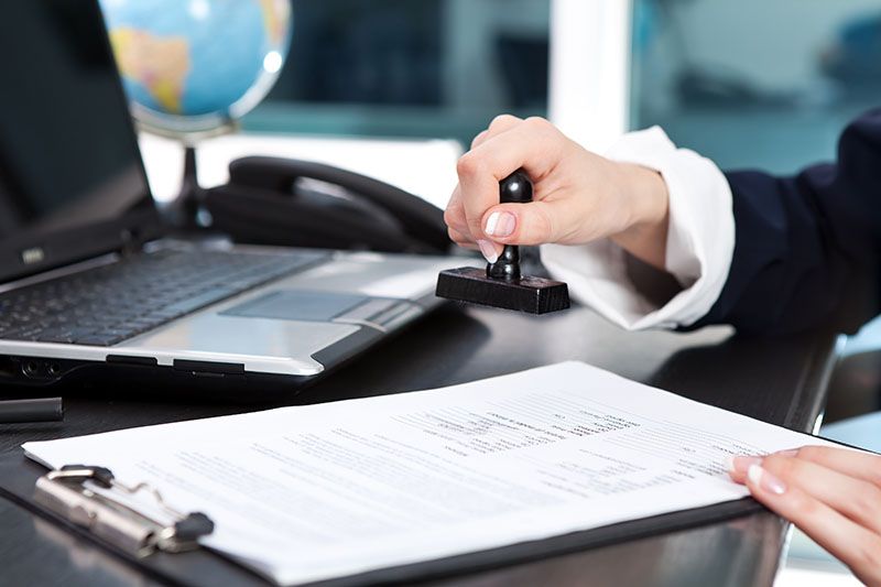 Learning Center 4 A person stamps a document on a clipboard at a desk with a laptop, phone, and globe in the background.