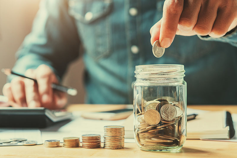 A person in a denim shirt drops a coin into a glass jar filled with coins. Stacks of coins, a notebook, and a hand using a calculator are visible on the table in the background.