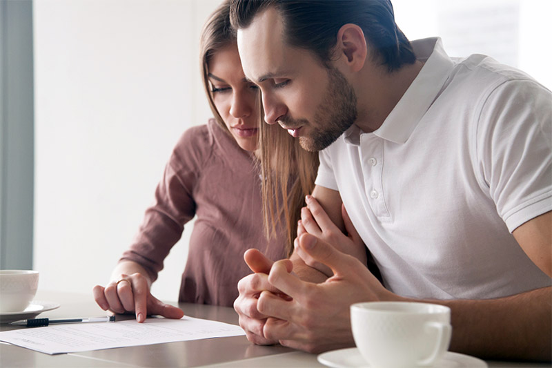 A couple sits closely together at a table, reading and discussing a document. One person points at the paper with a pen, while both look focused and serious. Two cups are on the table in front of them.