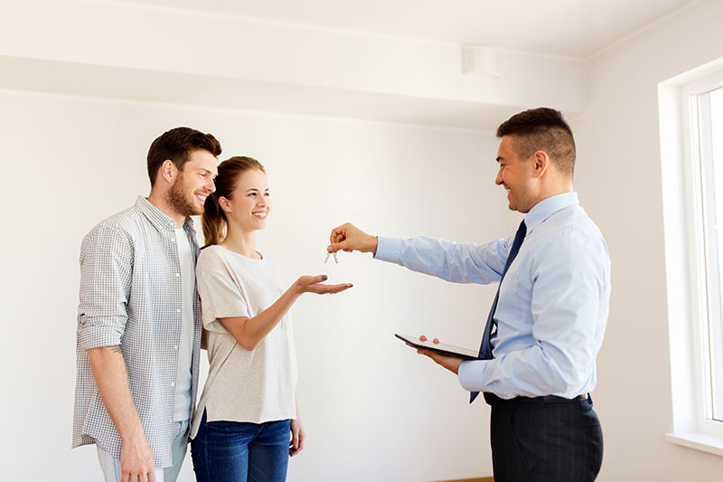 Home 162 A man in a suit hands keys to a smiling couple inside a bright, empty room, possibly signifying the purchase or rental of a new home. The woman reaches out to accept the keys.