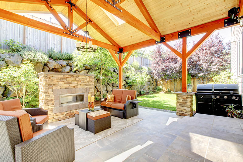 Home 161 Covered patio with wooden beams, wicker furniture, a stone fireplace, and a barbecue grill. Lush greenery and shrubs surround the outdoor space, creating a cozy and inviting backyard setting.