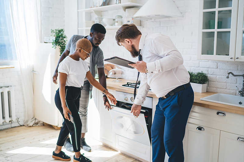 Home 184 A man in a suit shows a couple an oven in a modern white kitchen. The couple looks closely at the appliance while the man holds a clipboard and gestures toward the open oven door.