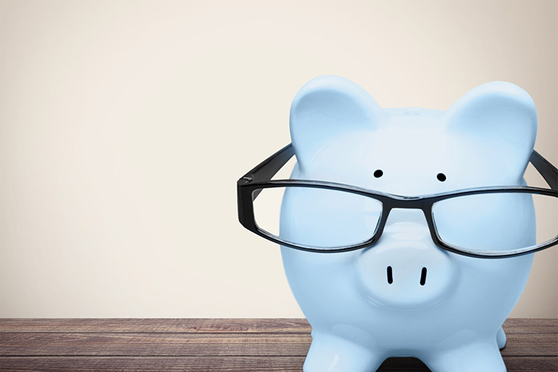 Home 173 A light blue piggy bank wearing black eyeglasses sits on a wooden surface against a plain, beige background.