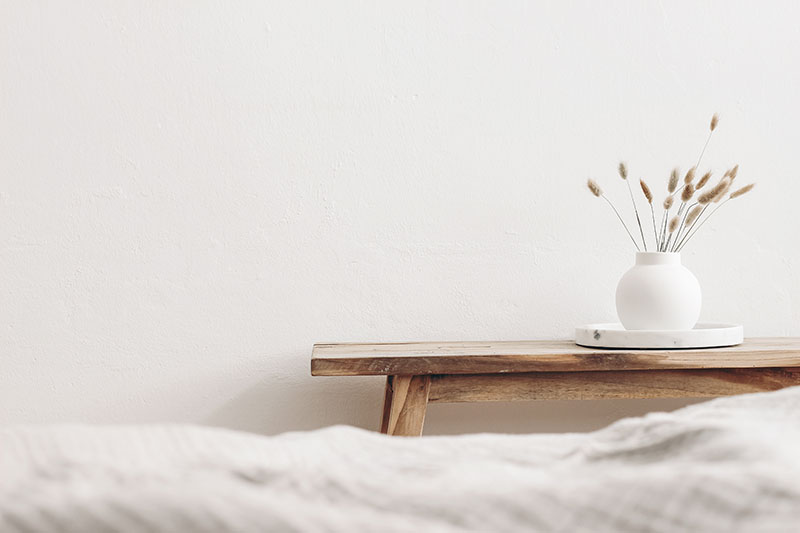 Home 190 A minimalist scene with a wooden bench against a white wall, holding a round white vase filled with dried grass on a white tray. The foreground shows the edge of a soft, light-colored blanket.