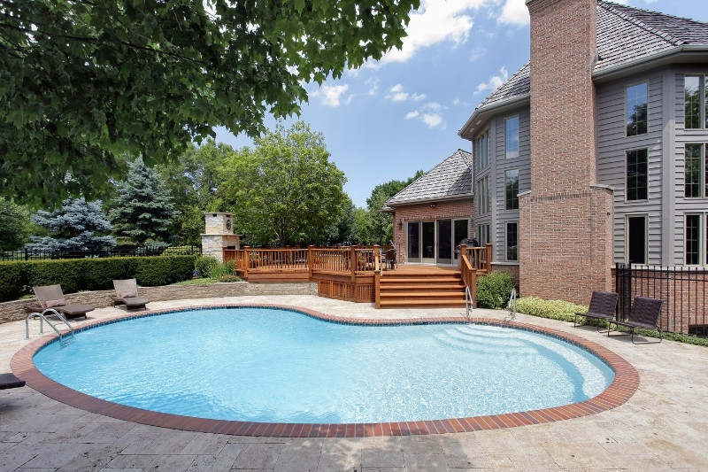 Home 149 A kidney-shaped swimming pool with a brick border in a backyard, surrounded by a patio, wooden deck, lounge chairs, and a large brick house with tall windows and a chimney. Lush greenery and trees are in the background.