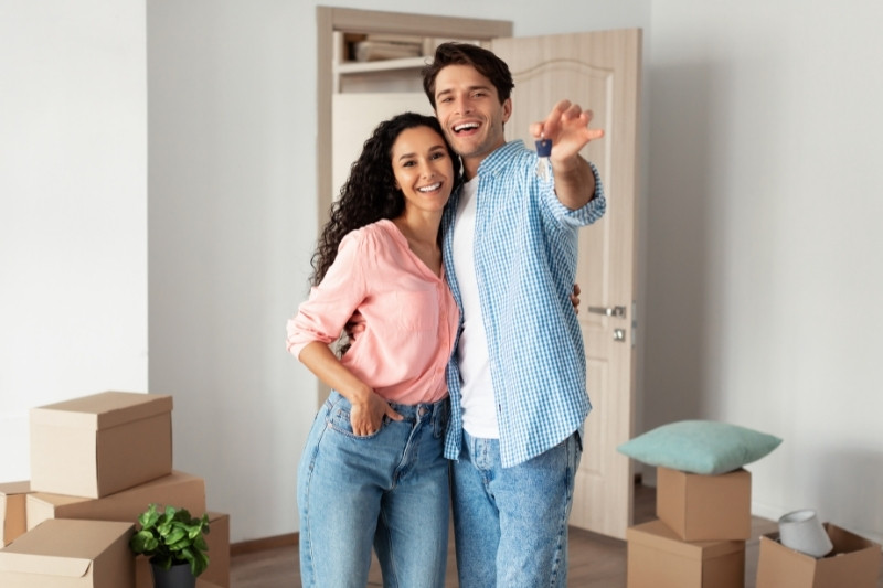 Home 150 A smiling couple stands close together in a room with moving boxes, with the man pointing playfully towards the camera. The room is bright and has an open door and a plant in the background.