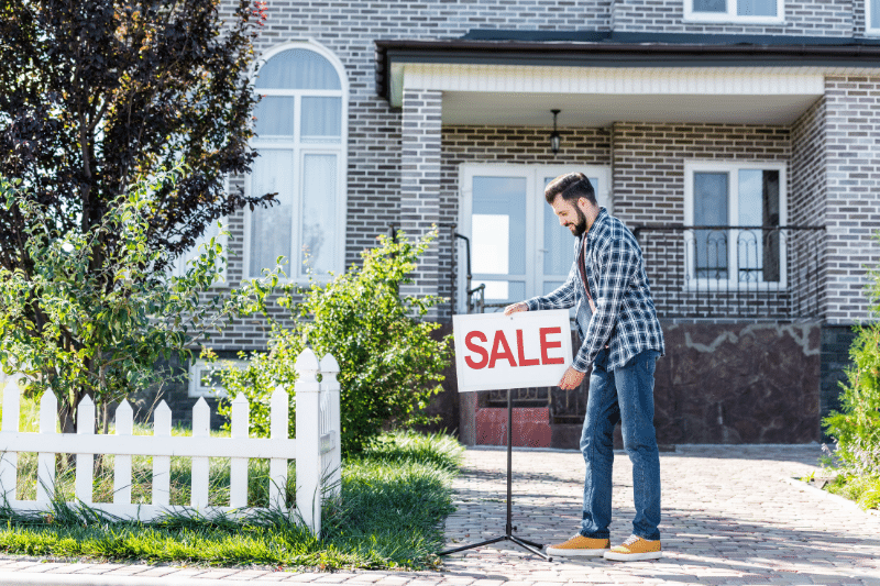 Home 148 A man places a SALE sign in front of a modern brick house with a white fence and green lawn.
