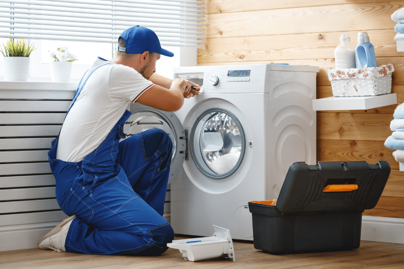 Home 145 A repairman in blue overalls kneels in front of a washing machine, using a tool to fix it. An open toolbox sits nearby in a bright laundry room with folded towels and cleaning supplies.