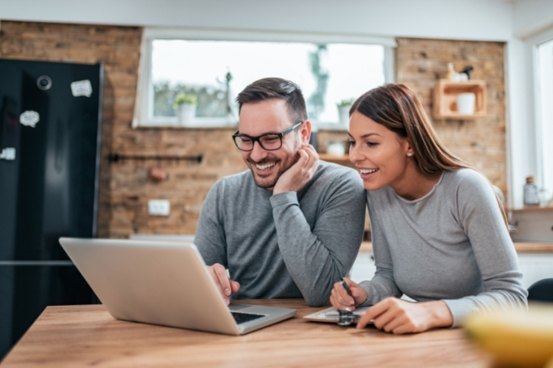 Home 138 A smiling couple sits at a kitchen table looking at a laptop. The man is wearing glasses and both are dressed casually. The woman holds a credit card and a notepad. The kitchen has a brick wall and a window in the background.