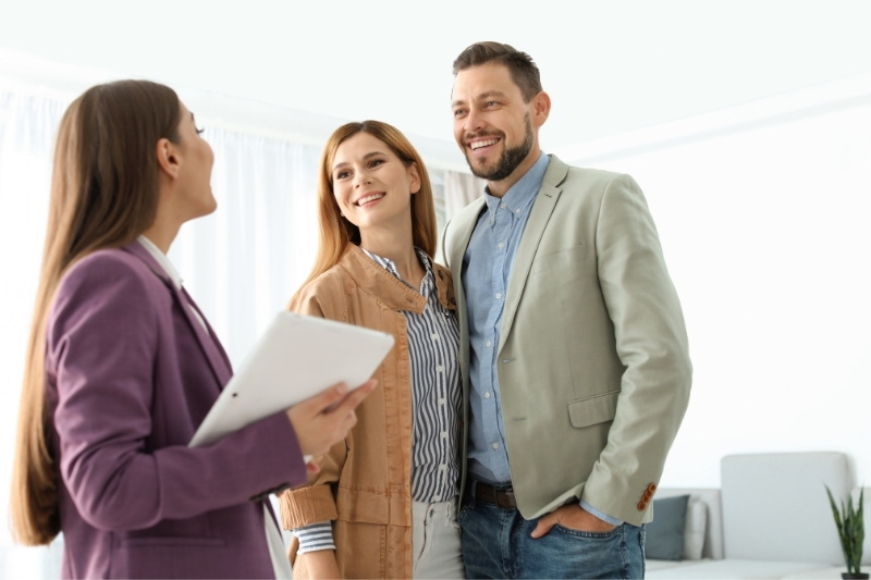 Home 139 A woman holding a tablet smiles while talking to a happy couple dressed in business casual attire, standing together in a bright, modern room.