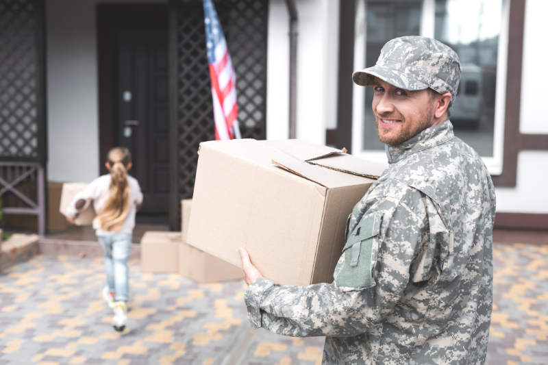 Home 133 A man in military uniform smiles while holding a cardboard box outside a house. An American flag is near the door, and a young girl carrying a box walks ahead of him toward the house.