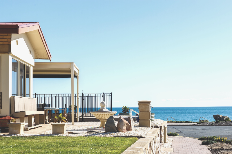 Home 131 A modern house with large windows and a patio overlooks a calm blue ocean. The yard has a fountain, rocks, and green grass, with a clear sky above and a paved walkway leading toward the water.