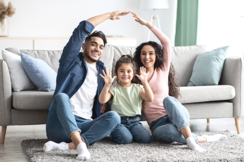 Home 132 A smiling family of three sits on a rug in front of a gray sofa. The adults make a roof shape with their arms over the child, who holds up both hands and smiles at the camera.